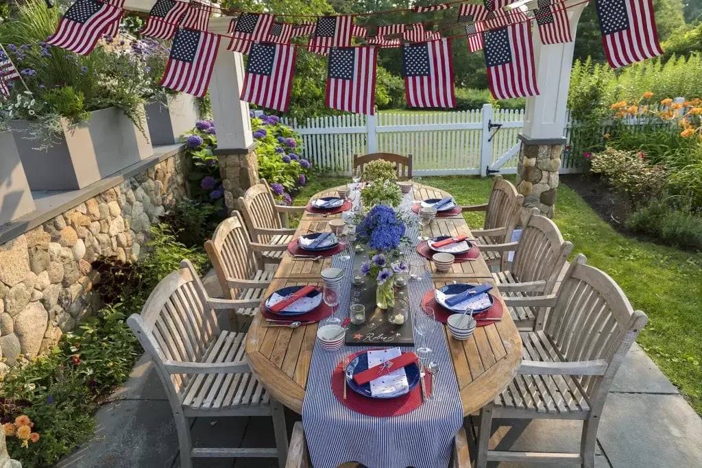 Large outdoor wooden table with 6 chairs with each place setting decorate in red, white, and blue for Labor Day, and with hanging rows of small American Flags fluttering above it.